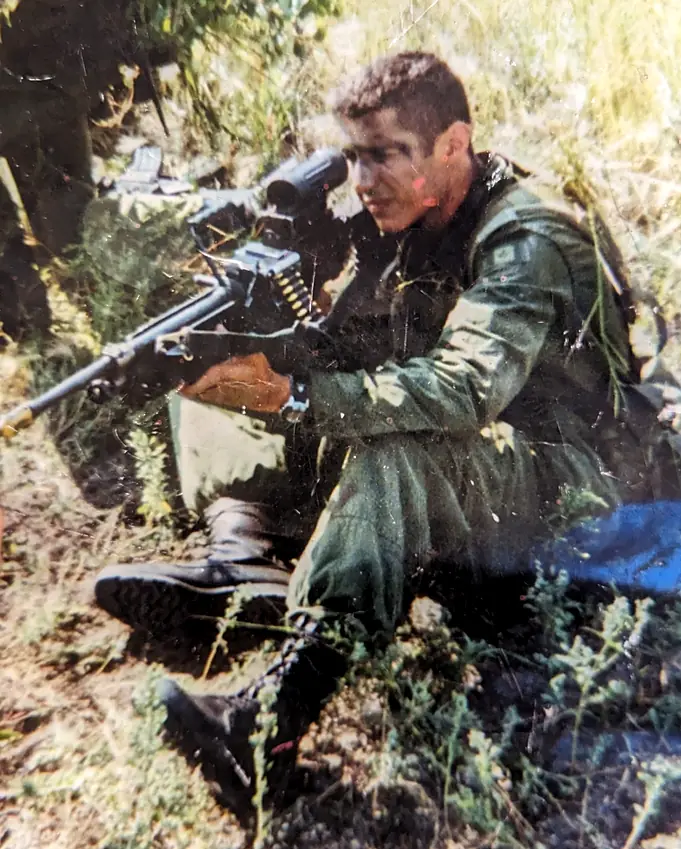 Charles Vincent in his late teens in combat uniform, seated in the field with a belt-fed machine gun across his lap, during his military service